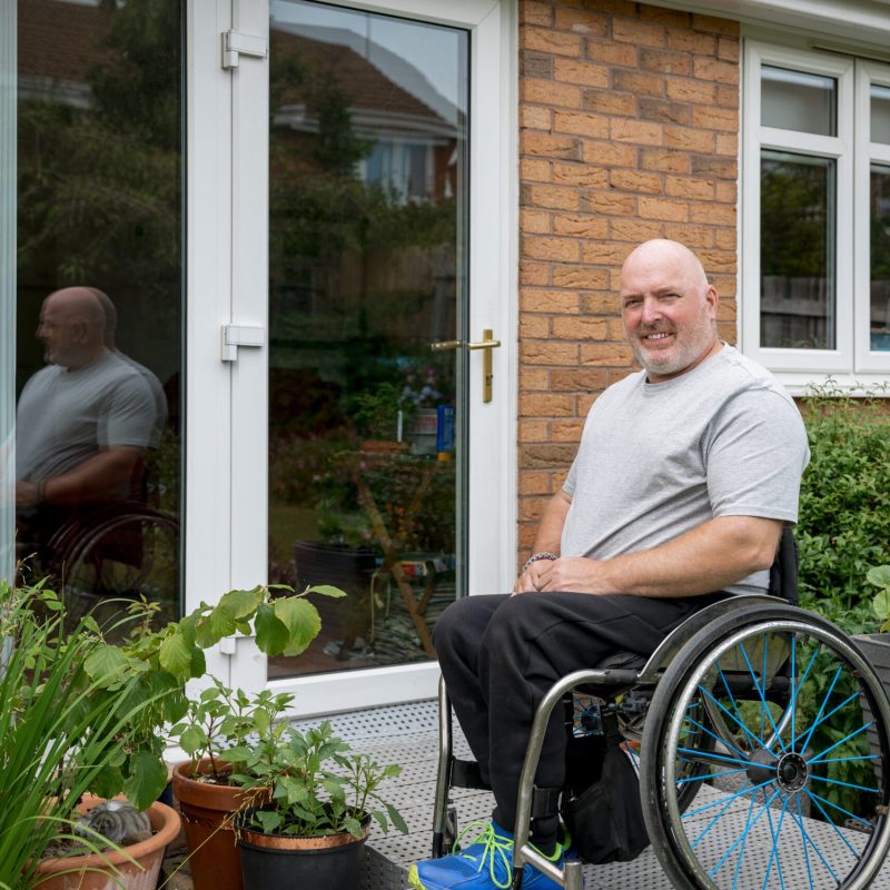 A portrait shot of a caucasian, mature man with paraplegia, outside of his home on a wheelchair ramp, looking and smiling at the camera.