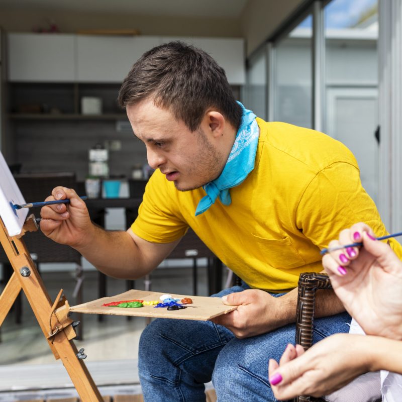 Teenager  with Down syndrome painting on canvas with his tutor on terrace above riverbank