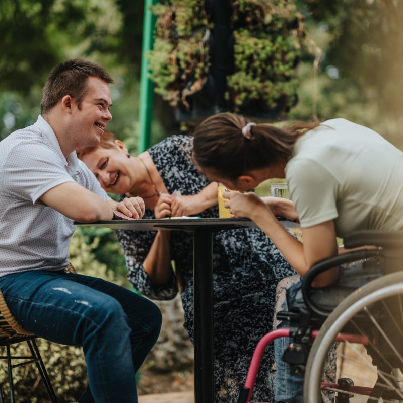 A group of friends, including a boy with Down syndrome and a girl in a wheelchair, share a laugh outdoors, highlighting friendship and inclusivity.