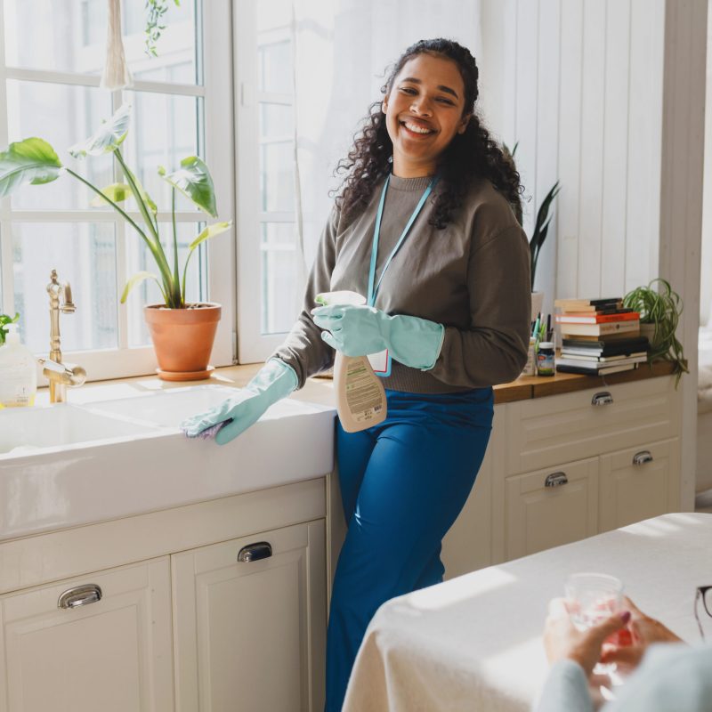 Selective focus on happy african american volunteer helping senior lady to clean house, standing in kitchen in raisin gloves washing sink with detergent, talking to elderly female sitting at table